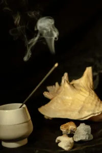 A burning incense stick in a white ceramic holder placed next to a large decorative seashell and stones.