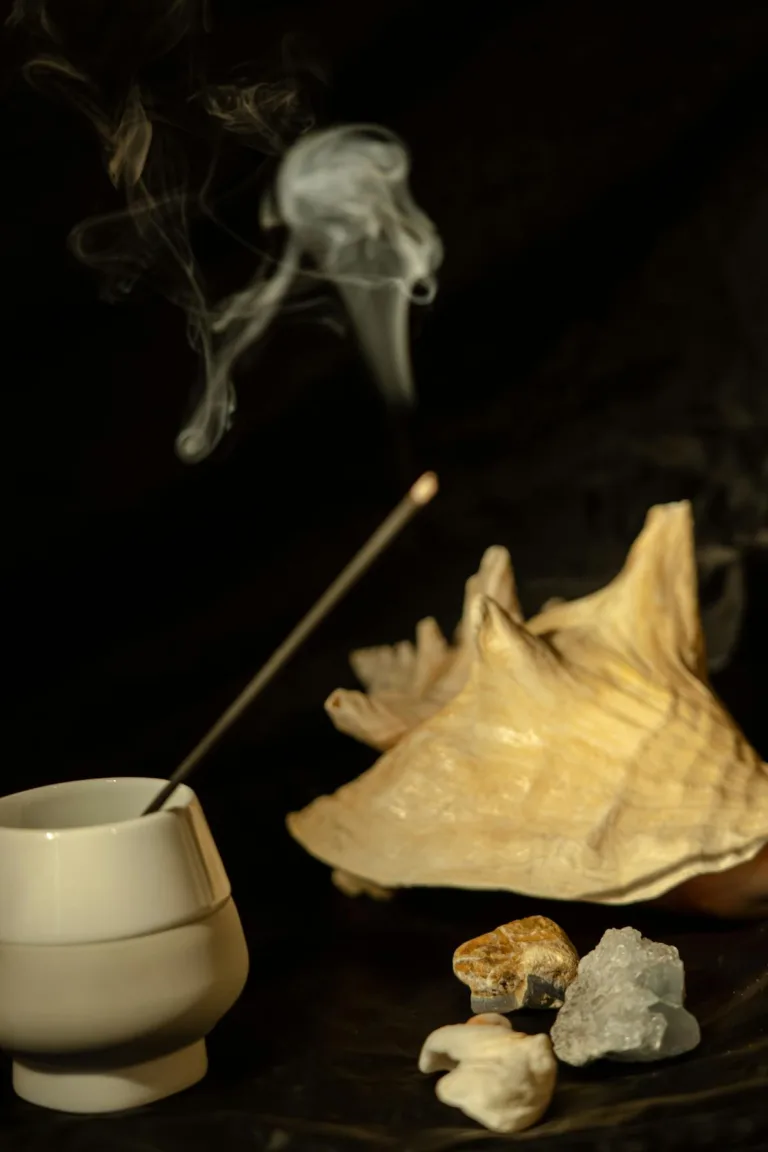 A burning incense stick in a white ceramic holder placed next to a large decorative seashell and stones.